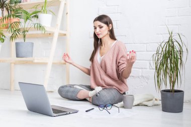 Genç mutlu kadın parlak bir dairede bir yoga poz yerde oturur ve bir dizüstü bilgisayar arkasında çalışır, çalışırken bir Freelancer kız meditasyon. kontrol stres