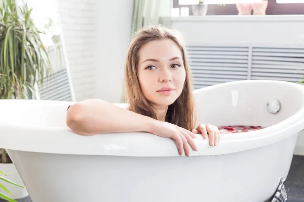 Een jong aantrekkelijk meisje neemt een badkamer met bloemblaadjes en ontspant tegen de achtergrond van een prachtig licht interieur. Spa-behandelingen voor schoonheid en gezondheid met huidverzorging — Stockfoto