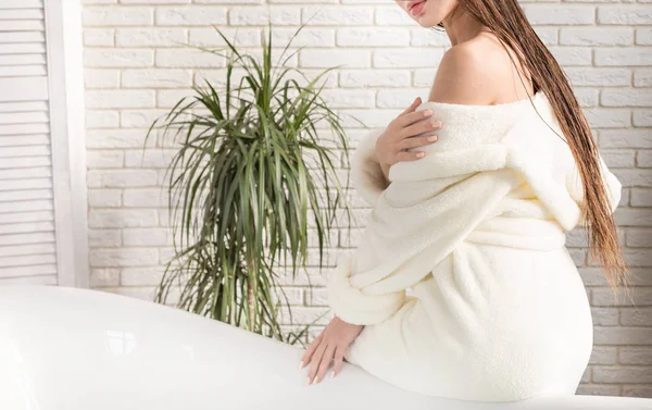 Die attraktive junge Frau im weißen Bademantel trägt Creme auf die Haut auf, während sie im Inneren des Badezimmers sitzt und morgendliche Behandlungen durchführt. Haut- und Körperpflege — Stockfoto