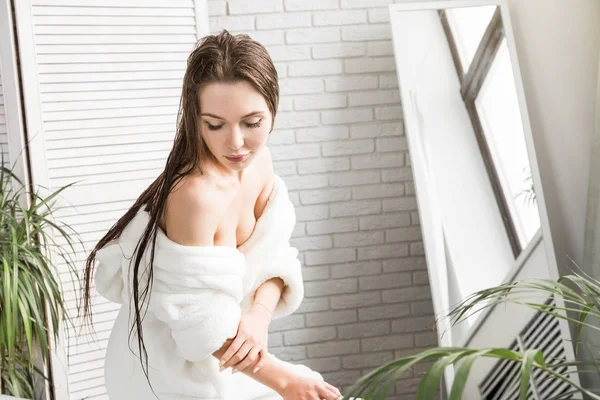 白いバスローブの魅力的な若い女の子は、バスルームの内部に座って朝の手順を実行しながら、肌にクリームを適用します。スキンケアとボディケア — ストック写真
