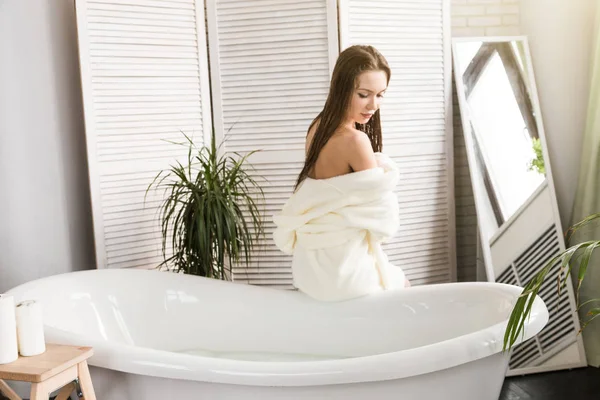 흰색 목욕용 가운을 입은 매력적 인 소녀는 욕실의 내부에 앉아 아침 절차를 밟는 동안 피부에 크림을 바르게 된다. 피부와 신체를 돌봄 — 스톡 사진