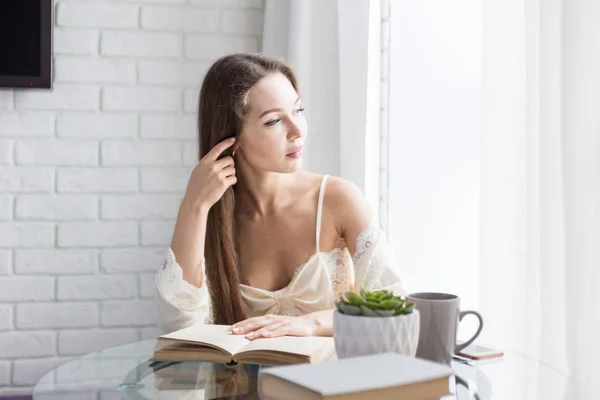 Menina bonita em uma camisola senta-se de manhã na mesa de vidro, olha pela janela e lê um livro — Fotografia de Stock