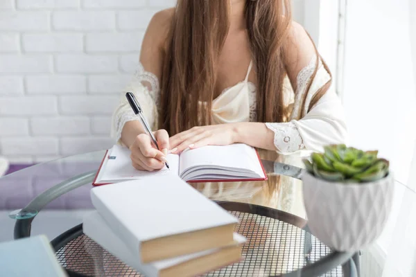 Een mooi jong meisje in een nightgown zit in de ochtend aan een glazen tafel en schrijft haar gedachten in een notebook. — Stockfoto