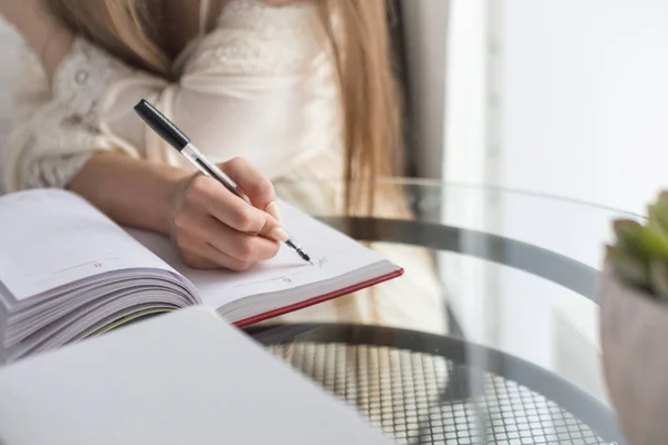 Una hermosa joven en un camisón está sentada en la mañana en una mesa de cristal y escribe sus pensamientos en un cuaderno . — Foto de Stock