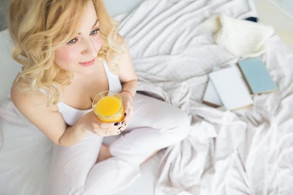 Aantrekkelijke blonde vrouw zit aan de rand van het bed thuis en houdt een kous met sinaasappelsap in haar hand. Ochtendstemming en kracht — Stockfoto