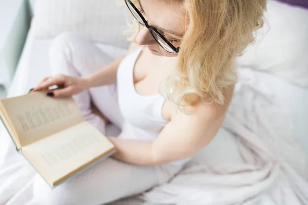 Atrakcyjna blondynka w okularach z czarną obręczą siedzi na krawędzi łóżka w domu z książką i czyta — Zdjęcie stockowe