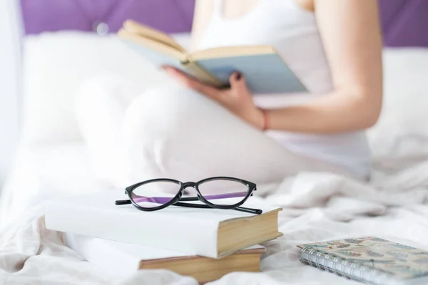 Aantrekkelijke vrouw blonde op de rand van het bed thuis met een boek en leest. Op de voorgrond een stapel boeken en brillen — Stockfoto