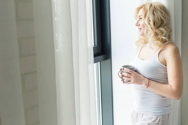 Aantrekkelijke blonde vrouw staat bij het raam met een kopje koffie of thee — Stockfoto