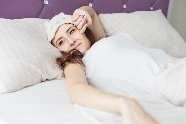 Een aantrekkelijke jonge brunette meisje met bruin haar wakker en slokjes terwijl geeuwen in haar bed in een slaapmasker — Stockfoto