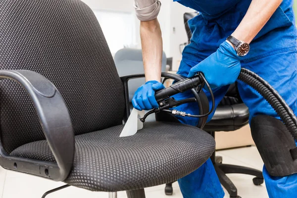 Jovem em vestuário de trabalho e luvas de borracha limpa a cadeira de escritório com equipamento profissional . — Fotografia de Stock