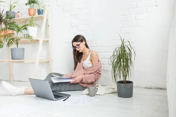 Mulher jovem senta-se no chão em um interior apartamento escandinavo com um laptop, estudando direito, menina freelance no trabalho, estudante de ensino à distância, emprego on-line, papelada e conceito de terceirização — Fotografia de Stock