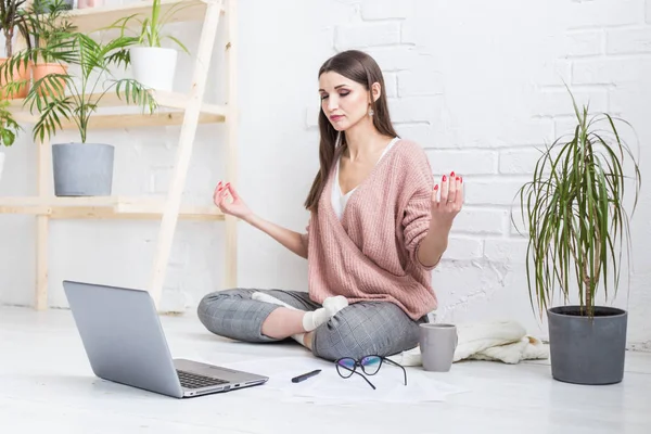Jovem mulher feliz senta-se no chão em uma pose de ioga em um apartamento brilhante e trabalha atrás de um laptop, uma menina freelancer medita enquanto trabalha. stress de controlo — Fotografia de Stock
