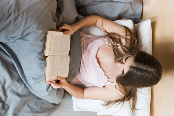 Jovem atraente em uma camisa de noite se senta na cama e lê um livro — Fotografia de Stock