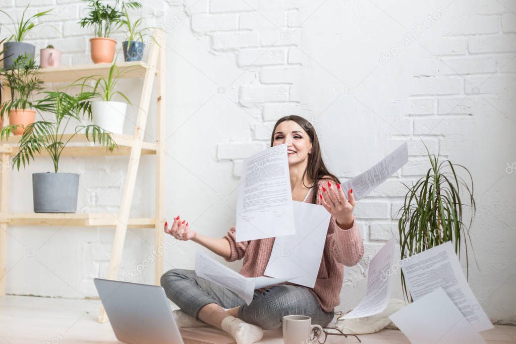 A young happy woman throws up a bunch of pieces of paper and documents sitting near the laptop, the freelance girl is happy and in a good mood. excitement and inspiration