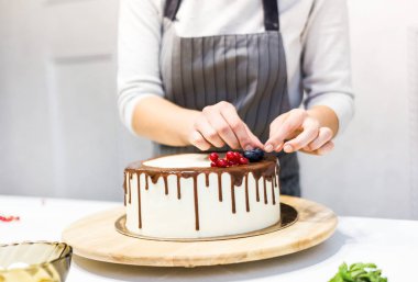 Tatlıcı çilek ile beyaz krem ve çikolata ile bir bisküvi kek dekorlar. Pasta beyaz bir masada ahşap bir stand üzerinde duruyor. Ev yapımı pasta kavramı, yemek pastaları.