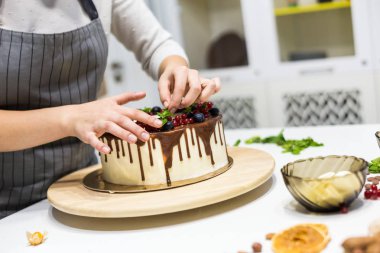 Tatlıcı çilek ile beyaz krem ve çikolata ile bir bisküvi kek dekorlar. Pasta beyaz bir masada ahşap bir stand üzerinde duruyor. Ev yapımı pasta kavramı, yemek pastaları.