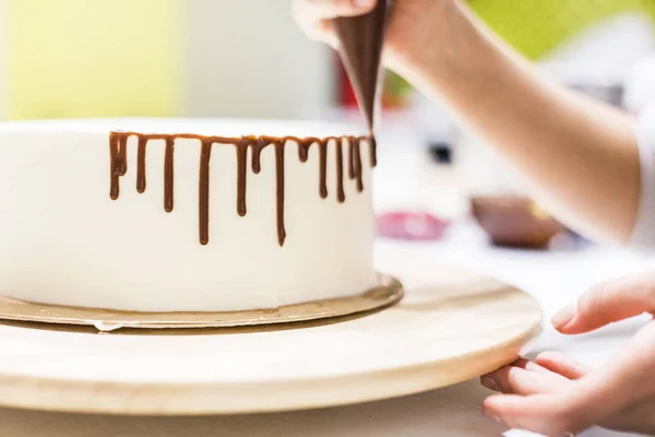 Um confeiteiro espreme chocolate líquido de um saco de pastelaria em um bolo de biscoito de creme branco em um suporte de madeira. O conceito de pastelaria caseira, bolos de cozinha . — Fotografia de Stock