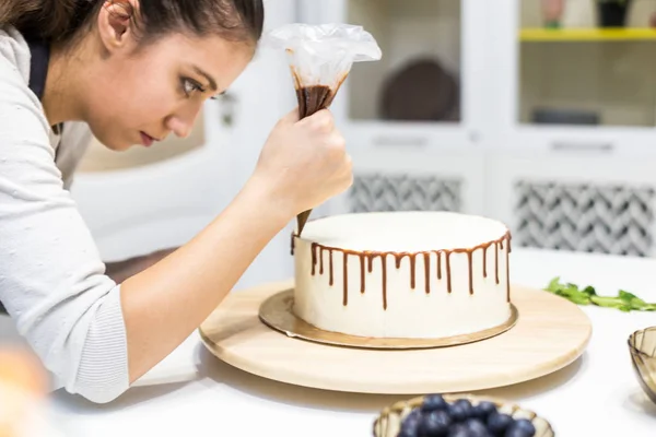 Een banketbakker knijpt vloeibare chocolade uit een gebakje zak op een witte room biscuit cake op een houten tribune. Het concept van zelfgemaakte gebak, koken cakes. — Stockfoto