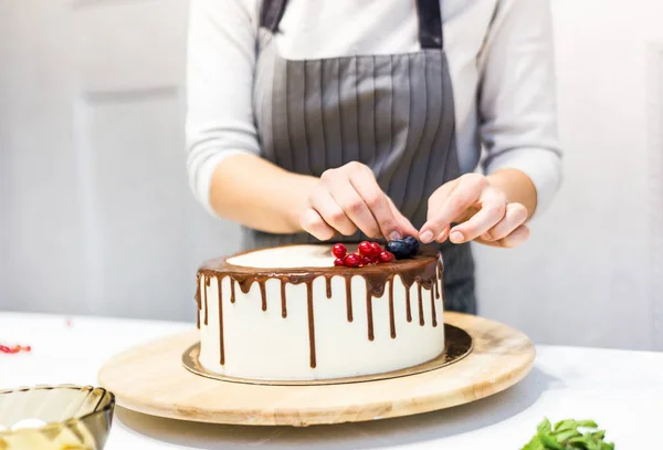 Confectioner dekorerar med bär en kak kaka med vit grädde och choklad. Kakan står på ett trä stativ på ett vitt bord. Konceptet med hembakat bak verk, matlagnings tårtor. — Stockfoto