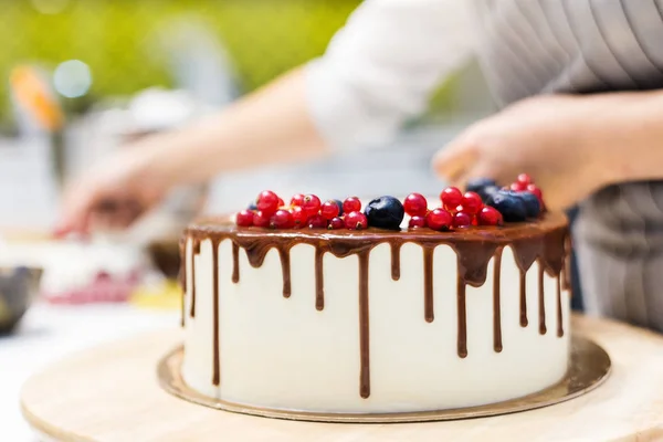 Il pasticcere decora con bacche una torta di biscotto con crema bianca e cioccolato. Cake si trova su un supporto di legno su un tavolo bianco. Il concetto di pasticceria fatta in casa, torte di cottura . — Foto Stock