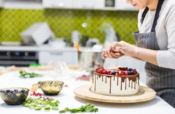 Cukrászda díszíti bogyókat keksz torta, fehér tejszín és csokoládé. A sütemény egy fából készült állványon áll egy fehér asztalon. A koncepció a házi készítésű péksütemények, főzősütemények. — Stock Fotó