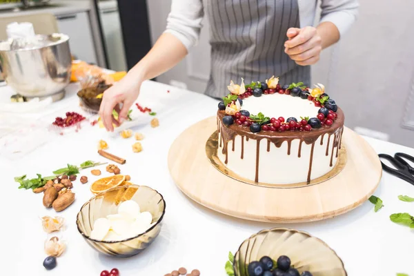 Confectioner dekorerar med bär en kak kaka med vit grädde och choklad. Kakan står på ett trä stativ på ett vitt bord. Konceptet med hembakat bak verk, matlagnings tårtor. — Stockfoto