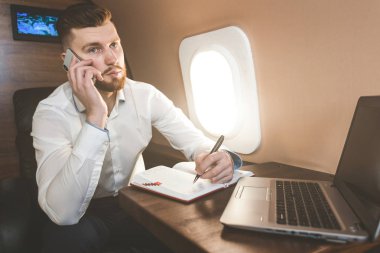 Genç, çekici ve başarılı bir iş adamı özel iş uçağında otururken telefonda konuşuyor ve çalışıyor.