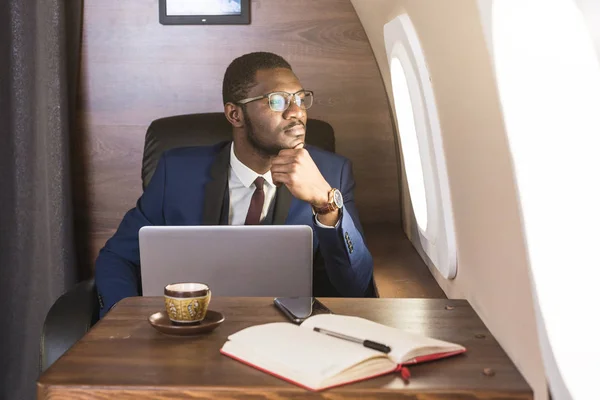 Atrakcyjny i udany African American biznesmen z okularami pracujących na laptopie siedząc na krześle jego prywatnego odrzutowca. — Zdjęcie stockowe