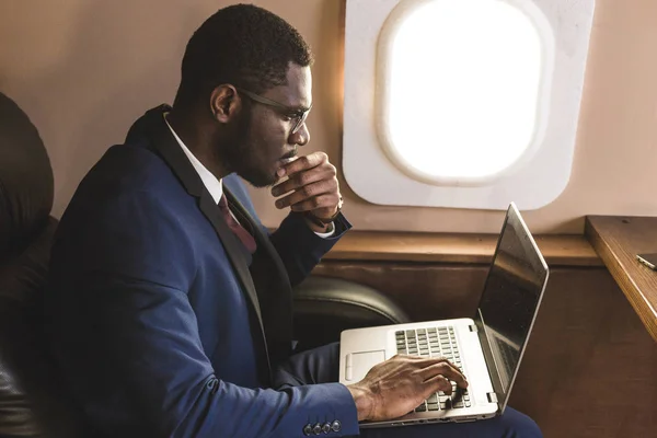 Atrakcyjny i udany African American biznesmen z okularami pracujących na laptopie siedząc na krześle jego prywatnego odrzutowca. — Zdjęcie stockowe