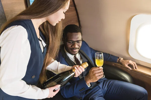 Молодой успешный афро-американский бизнесмен в очках и стюардесса показывает бутылку вина в салоне частного самолета. Сервис и полет первым классом . — стоковое фото