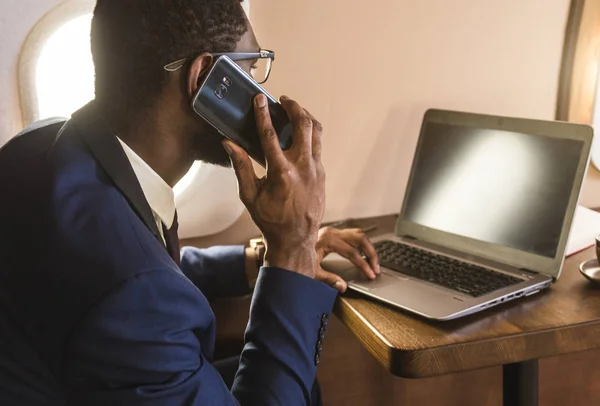 Młody atrakcyjny i udany African American biznesmen z glassies rozmowy na telefon i pracy siedząc na krześle jego prywatnej płaszczyźnie biznesowej — Zdjęcie stockowe