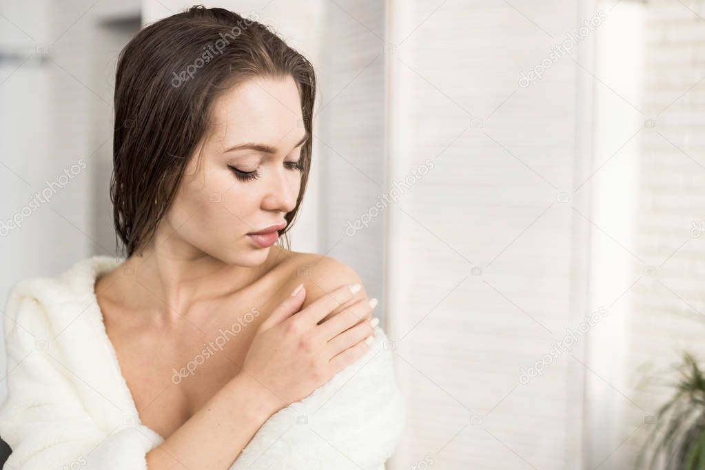 Attractive young girl in a white bathrobe applies cream to the skin while sitting in the interior of the bathroom and doing morning procedures. Skin and body care