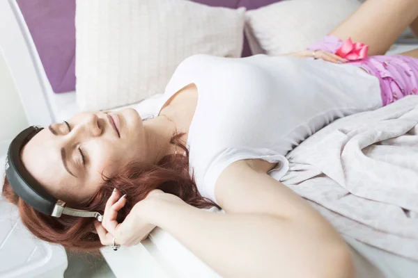 Een aantrekkelijke jonge brunette vrouw ligt in haar bed in de ochtend met haar hoofd hangen aan de rand met een koptelefoon en een smartphone en het luisteren naar muziek in een goed humeur — Stockfoto