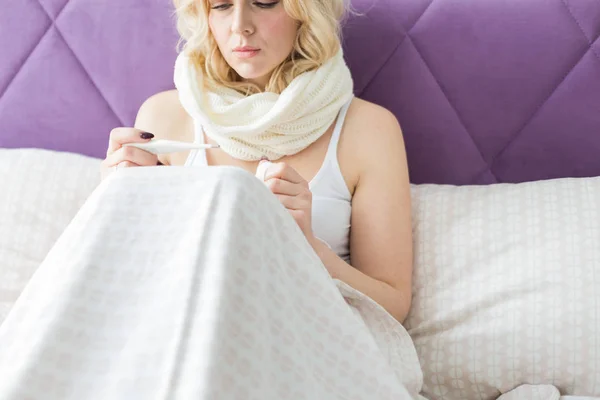 Mooie blonde vrouw zittend in bed met een thermometer en een sjaal gewikkeld rond de nek en een zakdoek. ziekte en gezondheid thuis — Stockfoto