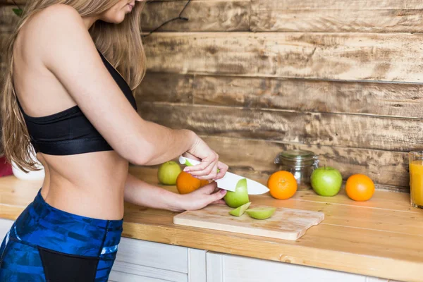 Attraktiv tjej i sportkläder förbereder en hälsosam frukost i köket innan träning, skära frukt och göra färsk juice. Hälsosam livsstil, fitness och beaut — Stockfoto