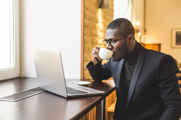 Joven hombre de negocios guapo de piel oscura en un café trabajando detrás de una computadora portátil con una taza de té . —  Fotos de Stock