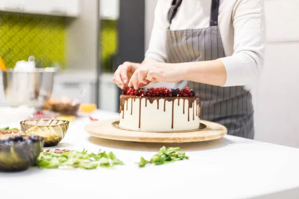Il pasticcere decora con bacche una torta di biscotto con crema bianca e cioccolato. Cake si trova su un supporto di legno su un tavolo bianco. Il concetto di pasticceria fatta in casa, torte di cottura . — Foto Stock