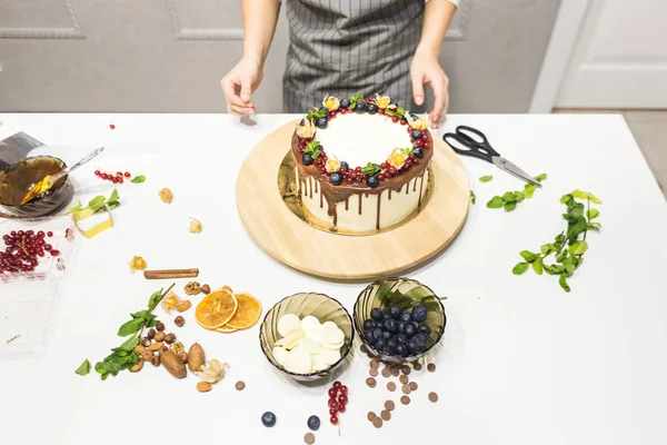 Il pasticcere decora con bacche una torta di biscotto con crema bianca e cioccolato. Cake si trova su un supporto di legno su un tavolo bianco. Il concetto di pasticceria fatta in casa, torte di cottura . — Foto Stock