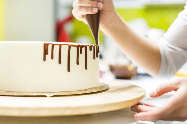 A confectioner squeezes liquid chocolate from a pastry bag onto a white cream biscuit cake on a wooden stand. The concept of homemade pastry, cooking cakes. — Stock Photo, Image