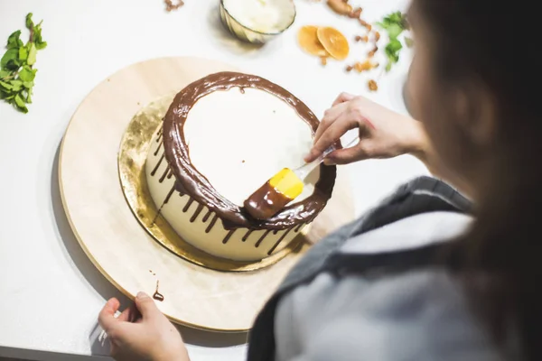 The confectioner decorates liquid chocolate biscuit cake with white cream, standing on a wooden stand. The concept of homemade pastry, cooking cakes.
