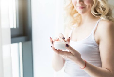 Güzel bir sarışın kadın elinde krem bir kavanoz tutar ve vücuduna uygular. Nemlendirici ile cilt bakımı