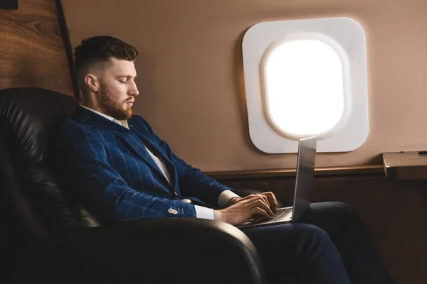 Atractivo y exitoso hombre de negocios que trabaja en una computadora portátil mientras está sentado en una silla de su jet privado . — Foto de Stock