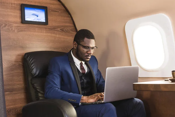 Vonzó és sikeres afro-amerikai üzletember a szemüveg dolgozik egy laptop ülve a szék az ő magángéppel. — Stock Fotó