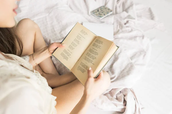Aantrekkelijk jong meisje in een nightgown zit in bed en leest een boek — Stockfoto