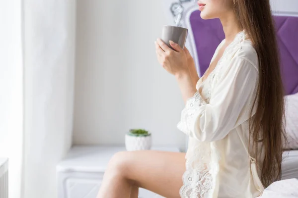 Приваблива молода дівчина в нічному одязі сидить на краю ліжка біля вікна і п'є каву з чашки — стокове фото