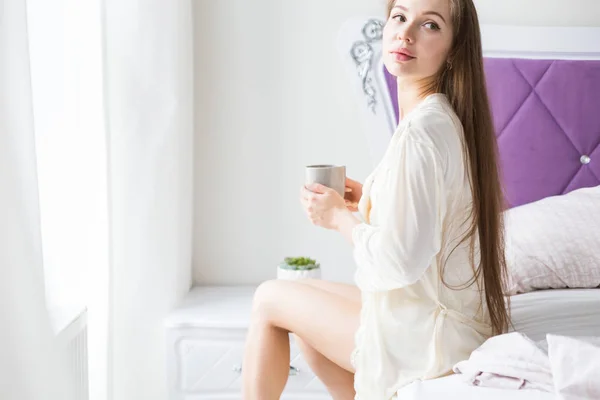 Atraktivní mladá dívka v nočních nojích sedí na kraji postele u okna a pije kávu z džbánu — Stock fotografie