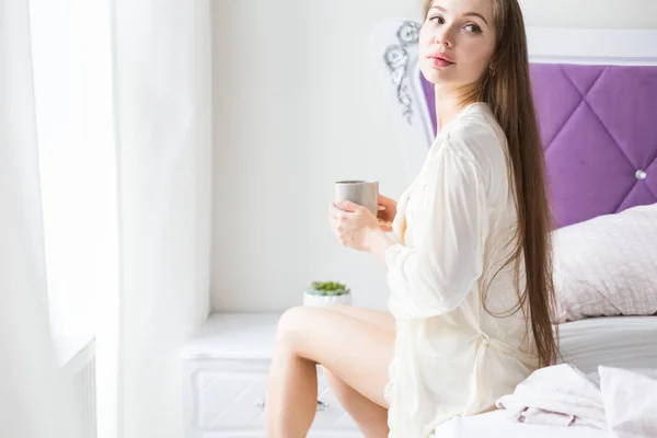 Atractiva joven en ropa de dormir se sienta en el borde de la cama junto a la ventana y bebe café de una taza — Foto de Stock