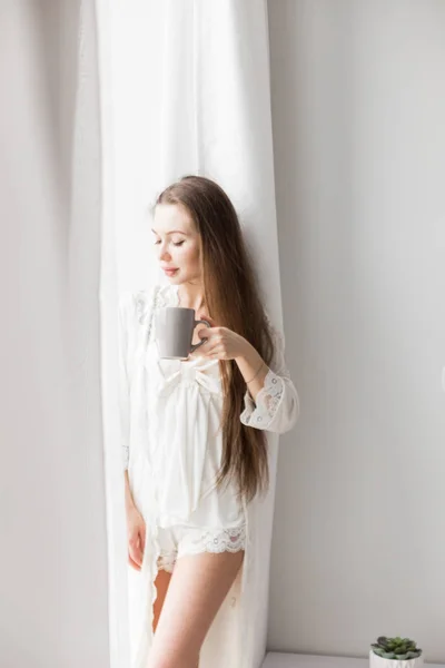 Attraktives junges Mädchen in Nachthemd, das mit einem Becher Kaffee am Fenster steht — Stockfoto
