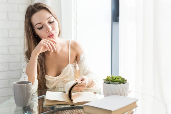Menina bonita em uma camisola senta-se de manhã na mesa de vidro, olha pela janela e lê um livro — Fotografia de Stock