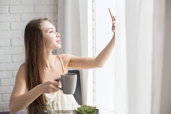 Uma menina bonita em uma camisa de noite senta-se de manhã em uma mesa de vidro, bebe café e tira uma selfie no telefone inicial . — Fotografia de Stock